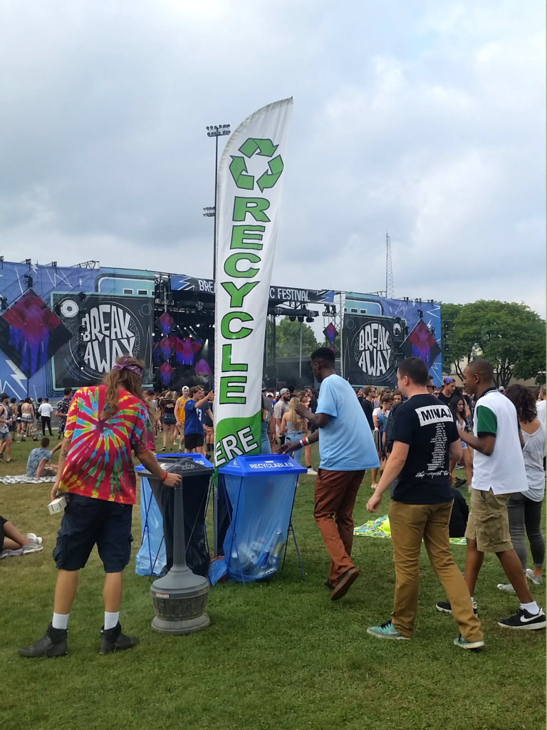 Photo of young festival attendees recycling in clear stream bin. Large Recycling sign takes up the center of the scene. Breakaway Michigan stage in the background.